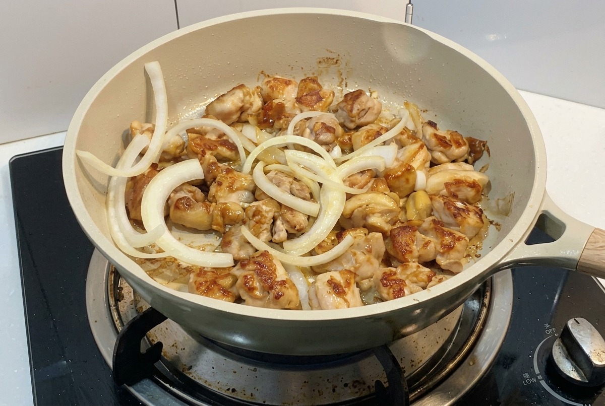 Sliced white onion rings being added to a pan of browning chicken pieces on a stove.