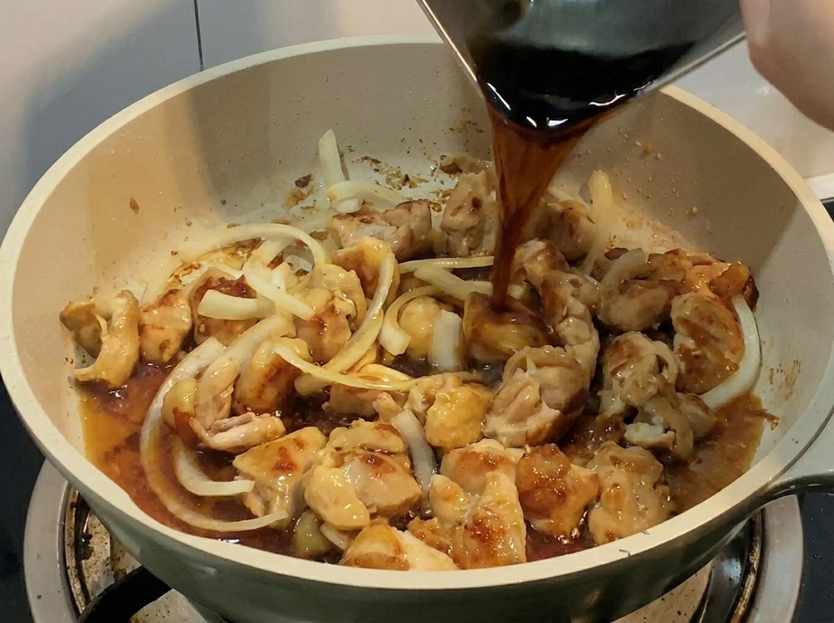 A dark, savory liquid being poured from a metal container into a frying pan filled with chicken and onions.