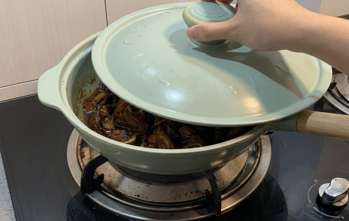 A hand placing a light blue lid onto a light green frying pan containing chicken and onions simmering in a dark sauce.