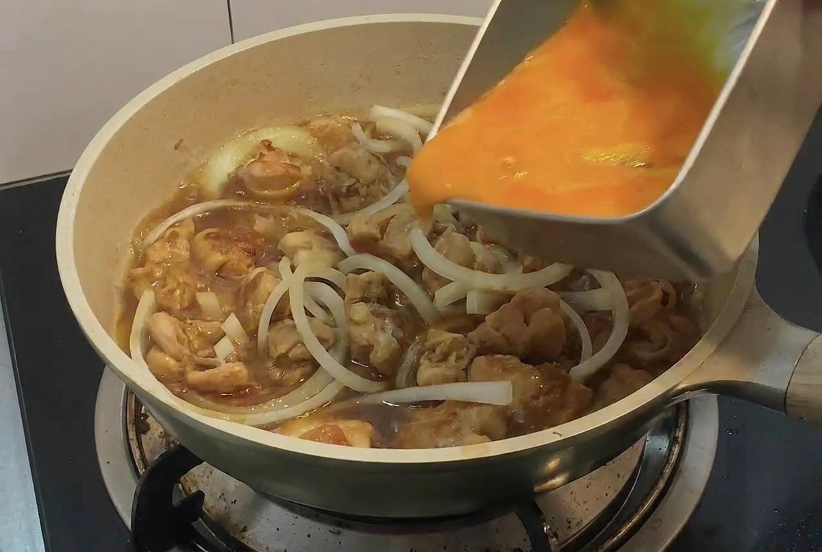 A hand pouring beaten egg from a rectangular metal tray into a pan with chicken and onion slices.