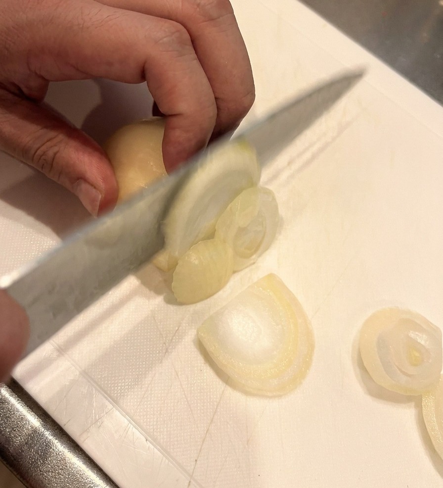 Ein scharfes Messer schneidet eine weiße Zwiebel in feine Streifen auf einem weißen Schneidebrett, gehalten von einer Hand.