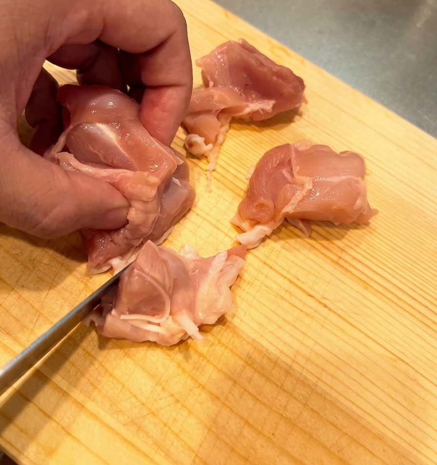 Rohes Hähnchenschenkelfleisch wird auf einem hellen Holzbrett in mundgerechte Stücke geschnitten.