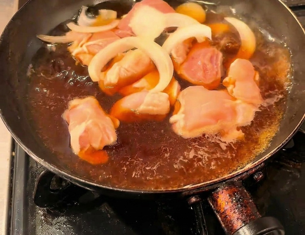 Hähnchenstücke und Zwiebelstreifen köcheln in einer dunklen Sojasaucenbrühe in einer schwarzen Pfanne.