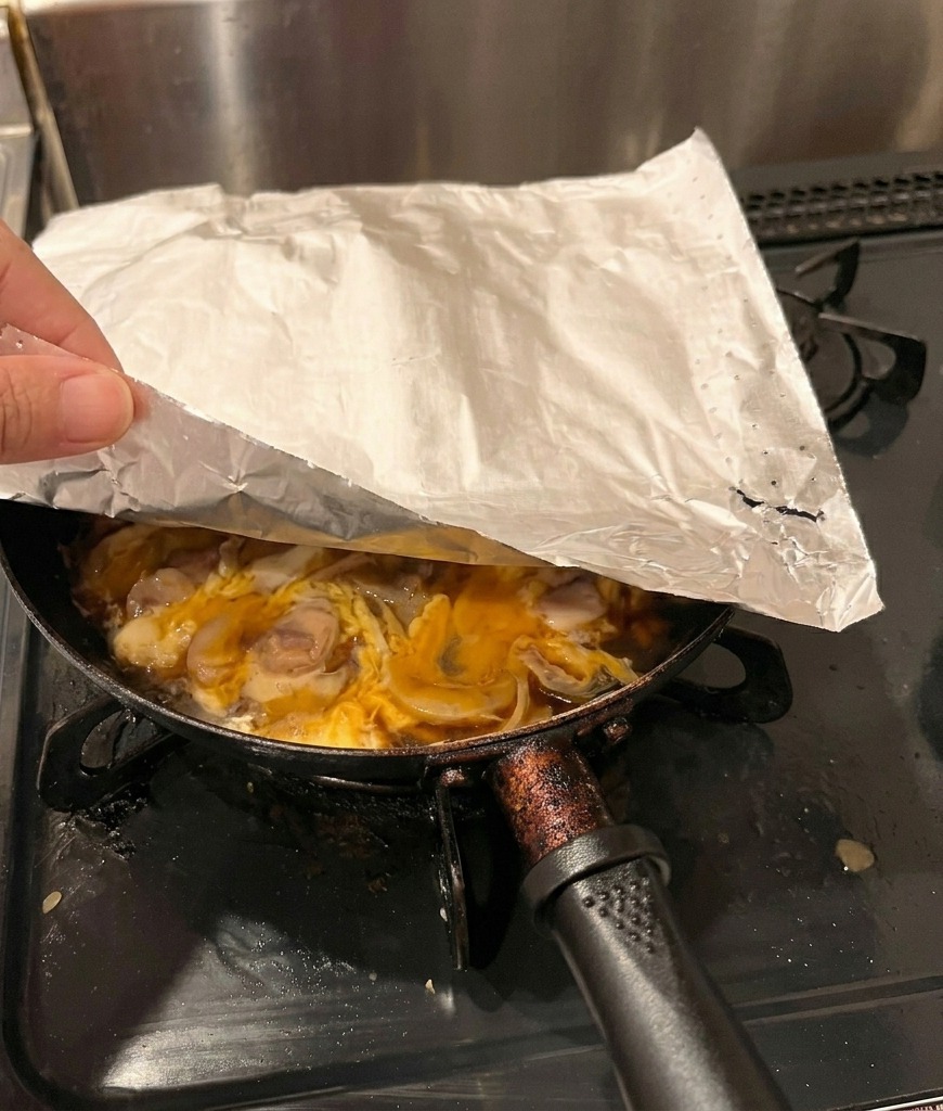 Eine Hand legt ein rundes Stück Backpapier über eine Pfanne mit Hähnchen und teilweise gestocktem Ei.