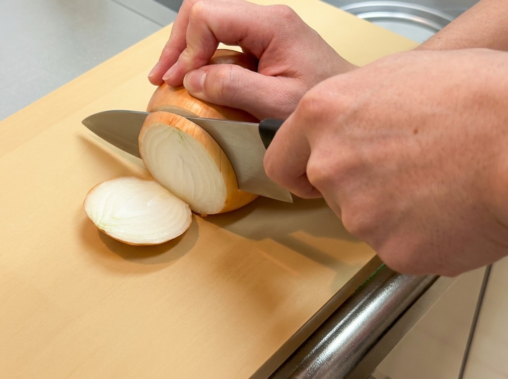 Eine Person halbiert eine geschälte gelbe Zwiebel der Länge nach auf einem hellen Schneidebrett.