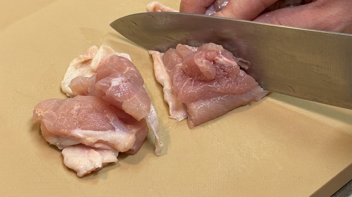 Rohes Fleisch von der Hähnchenkeule wird mit einem großen Küchenmesser in gleichmäßige, mundgerechte Stücke geschnitten.