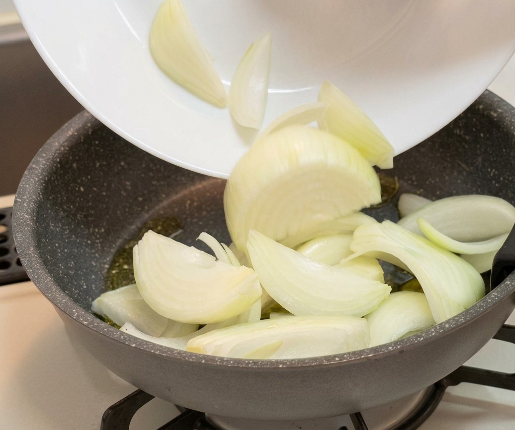Geschnittene weiße Zwiebelspalten werden von einem weißen Teller in eine graue, beschichtete Bratpfanne gegeben.