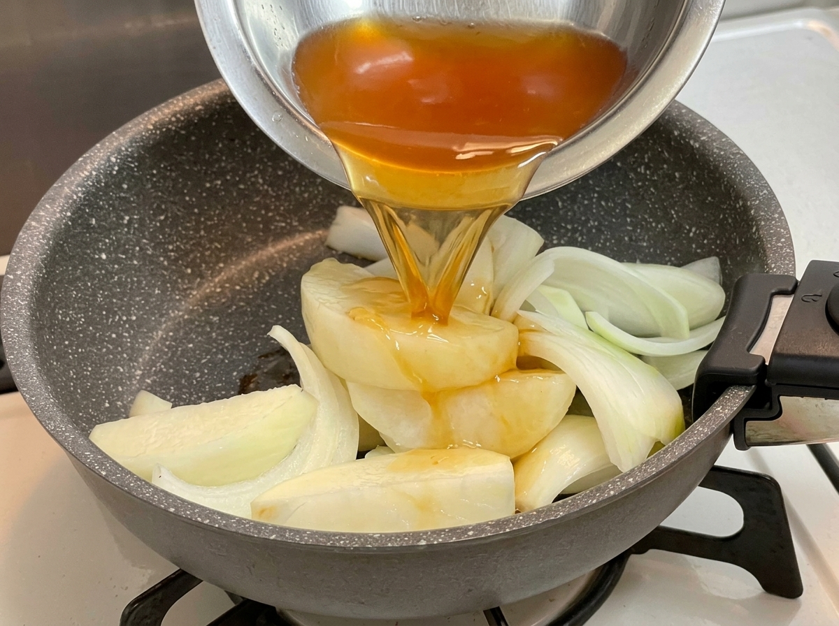 Dunkle Würzsauce wird aus einer Metallschüssel über die Zwiebelspalten in der Bratpfanne gegossen.