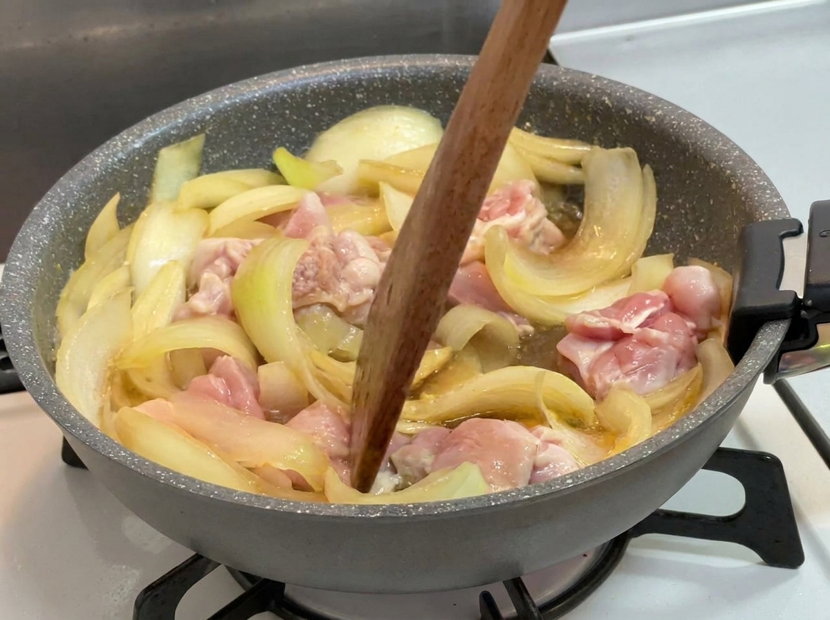Ein Holzwender rührt leicht gegartes Hähnchenfleisch und Zwiebeln in einer köchelnden, dunklen Flüssigkeit um.