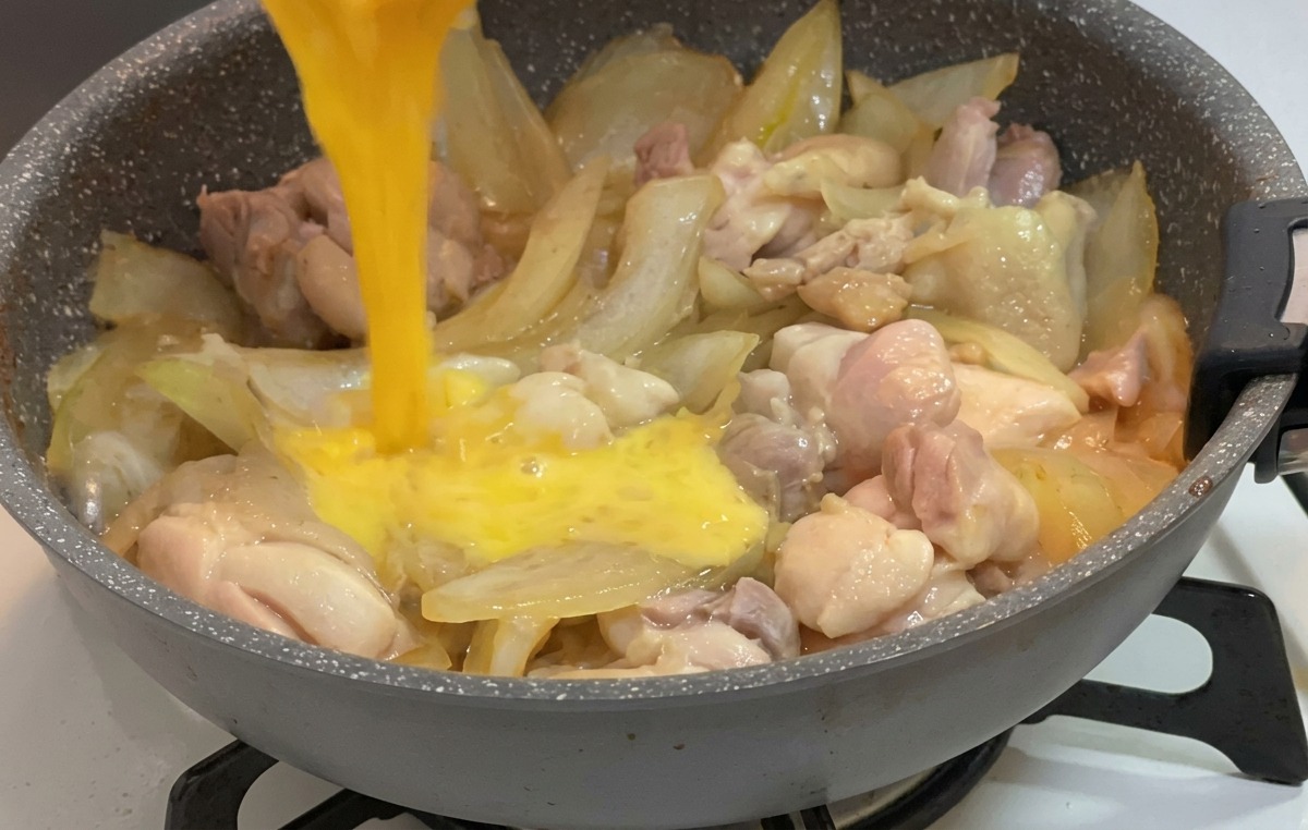 Verquirltes Ei wird aus einer kleinen Schüssel auf das in der Sauce köchelnde Hähnchen und die Zwiebeln gegossen.