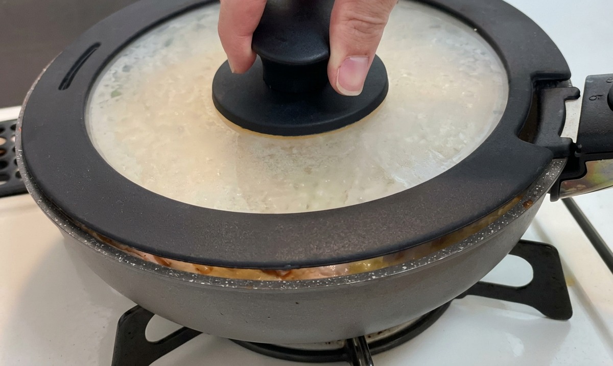 Eine Hand setzt einen Glasdeckel mit schwarzem Knauf auf eine Pfanne mit Hähnchen, Zwiebeln und Ei.