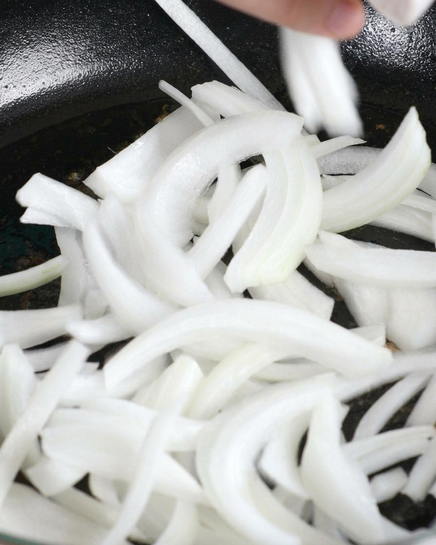 In Streifen geschnittene weiße Zwiebeln werden in die Pfanne mit dem verbliebenen Hähnchenfett gegeben.