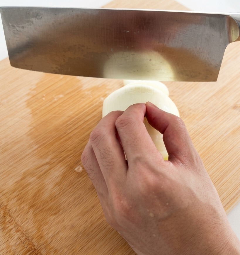 Eine Hand schneidet eine weiße Zwiebel mit einem großen Edelstahl-Hackmesser auf einem Bambus-Schneidebrett in feine Streifen.