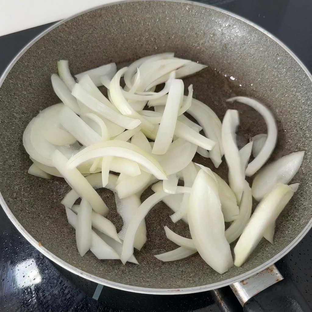 Fein geschnittene weiße Zwiebeln werden im verbliebenen Hähnchenfett in einer dunklen Pfanne gebraten, bis sie weich sind.