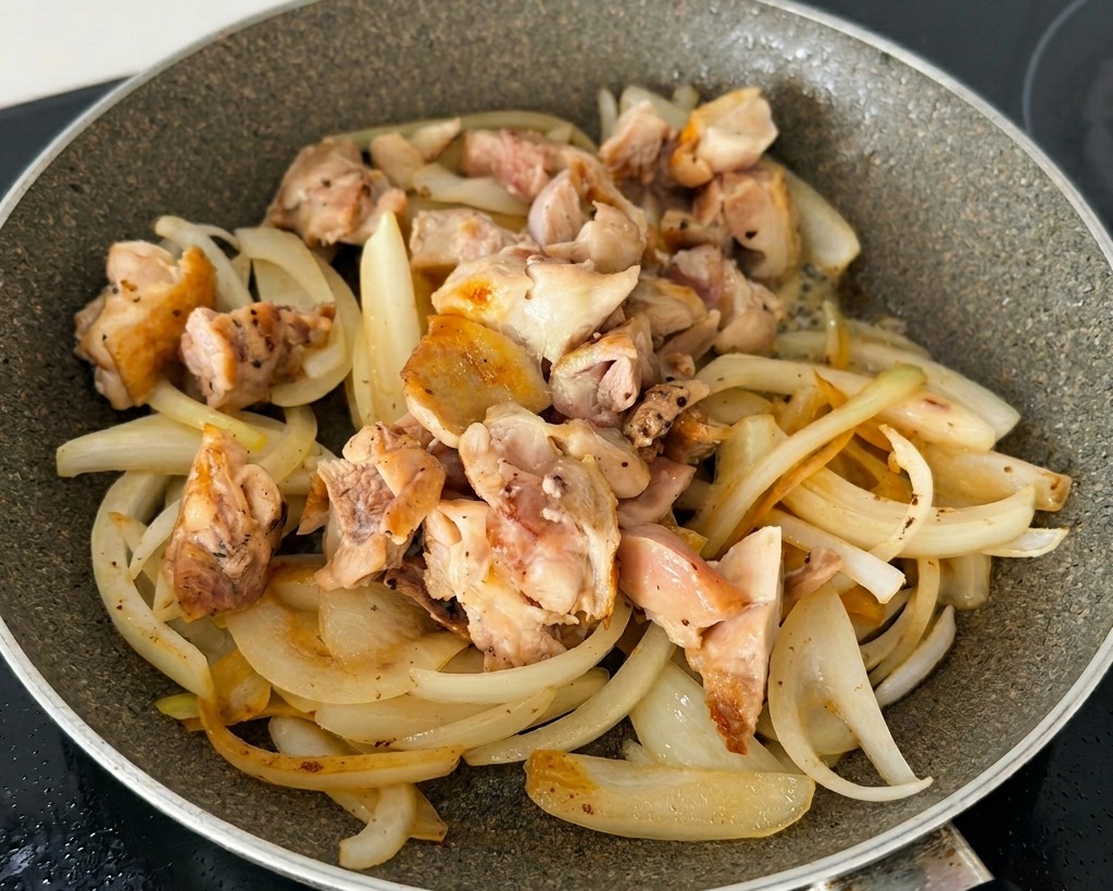 Angeschwitzte Zwiebelstreifen gemischt mit gebräunten Hähnchenstücken und Frühlingszwiebeln in einer beschichteten Bratpfanne.