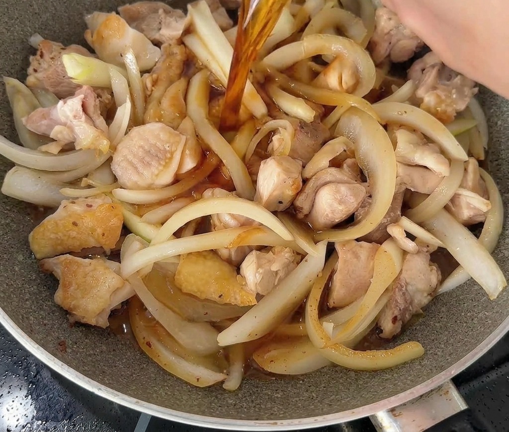 Eine dunkle, würzige Flüssigkeit wird aus einer weißen Schüssel in eine Pfanne mit Hähnchen und Zwiebelstreifen gegossen.