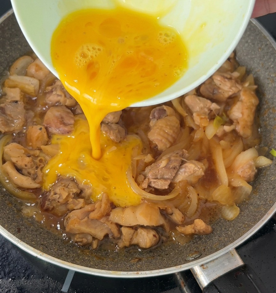Ein Strahl verquirlten Eies wird über das köchelnde Hähnchen und die Zwiebeln in einer Bratpfanne gegossen.