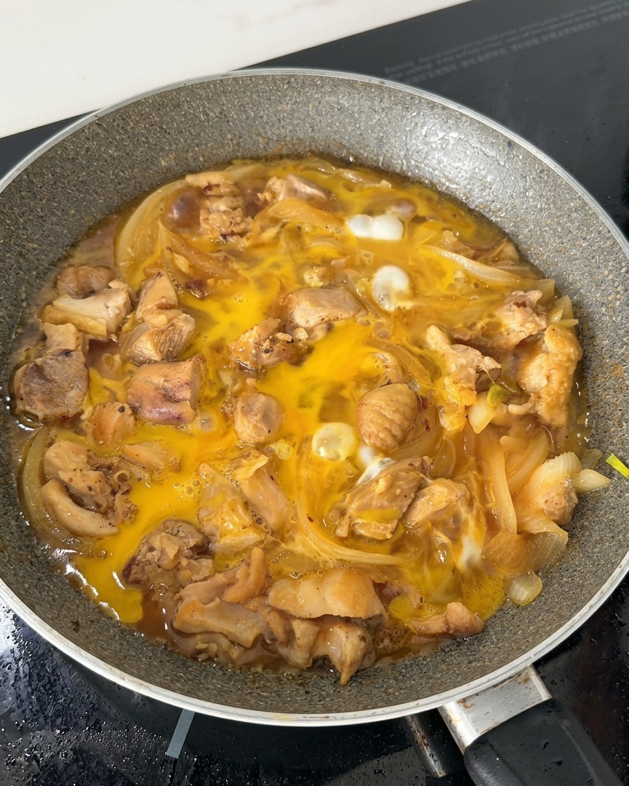 Die teilweise gestockte Eimischung blubbert in einer Pfanne mit Hähnchenstücken und Zwiebelstreifen in einer Brühe auf Sojabasis.