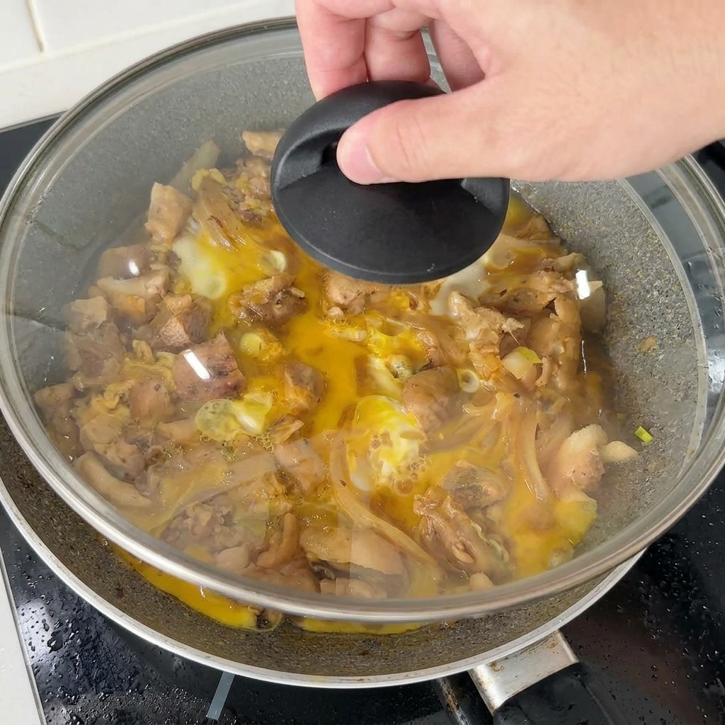 Eine Hand setzt einen Glasdeckel auf eine Pfanne mit Hähnchen und Eiern, um die zuletzt hinzugefügte Eimischung zu dämpfen.