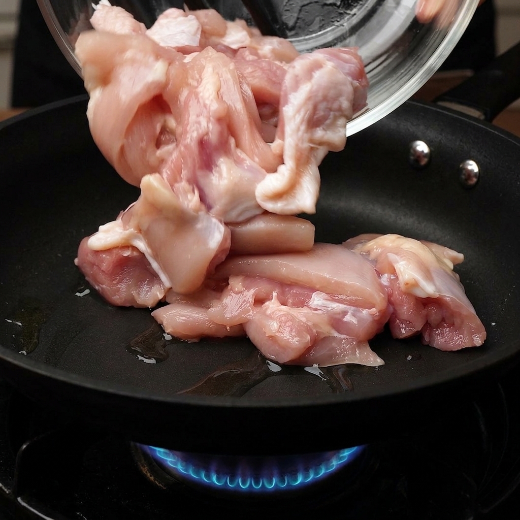 Rohe Hähnchenstücke, die aus einer Glasschale in eine heiße Bratpfanne gegeben werden, unter der eine blaue Gasflamme brennt.