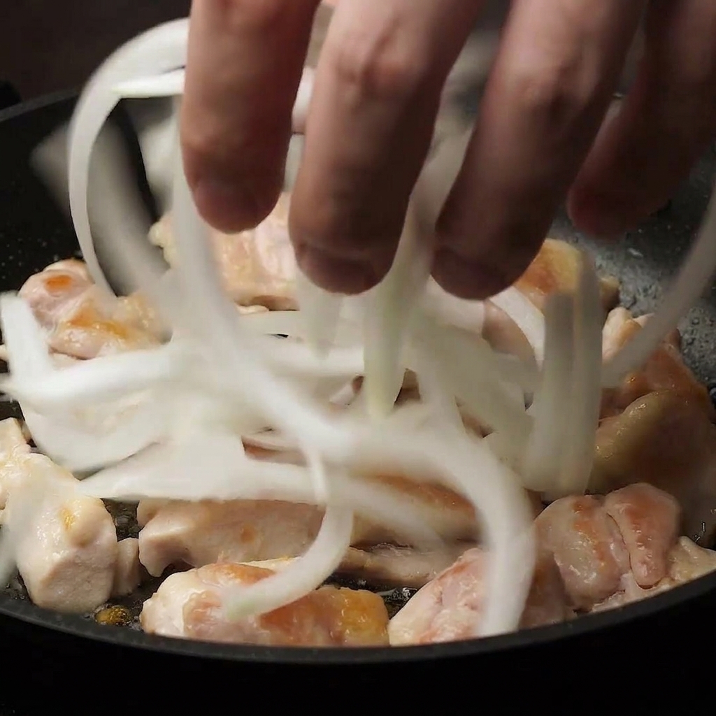 Eine Hand, die dünn geschnittene weiße Zwiebelstreifen in eine Pfanne zu den angebratenen Hähnchenstücken gibt.