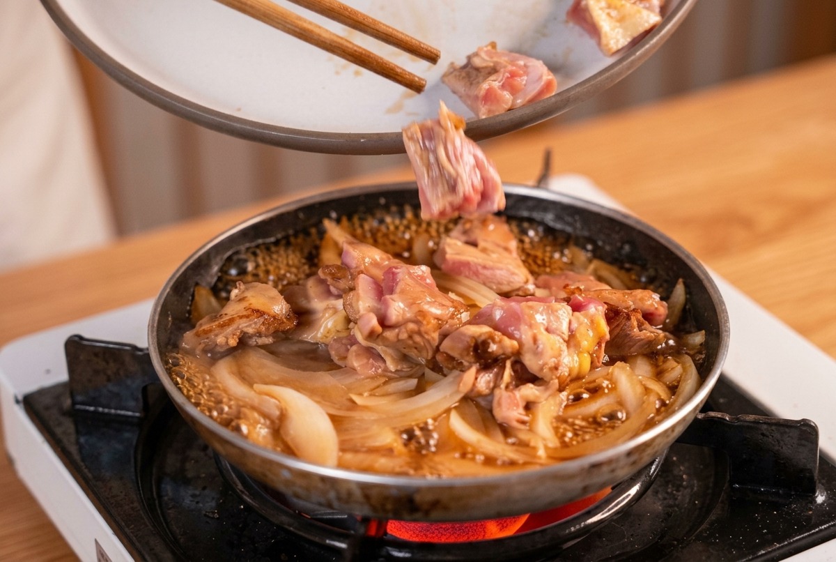 Angebratene Hähnchenstücke werden mit Stäbchen zurück in die Pfanne zu den köchelnden Zwiebeln und der Dashi-Brühe gelegt.