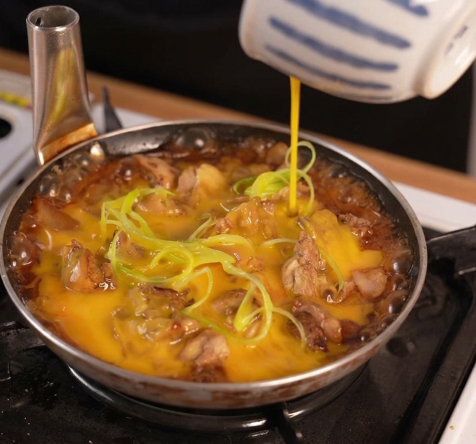 Verquirlte Eimasse wird aus einer gestreiften Schale über das Hähnchen und die Frühlingszwiebeln in die kochende Sauce gegossen.
