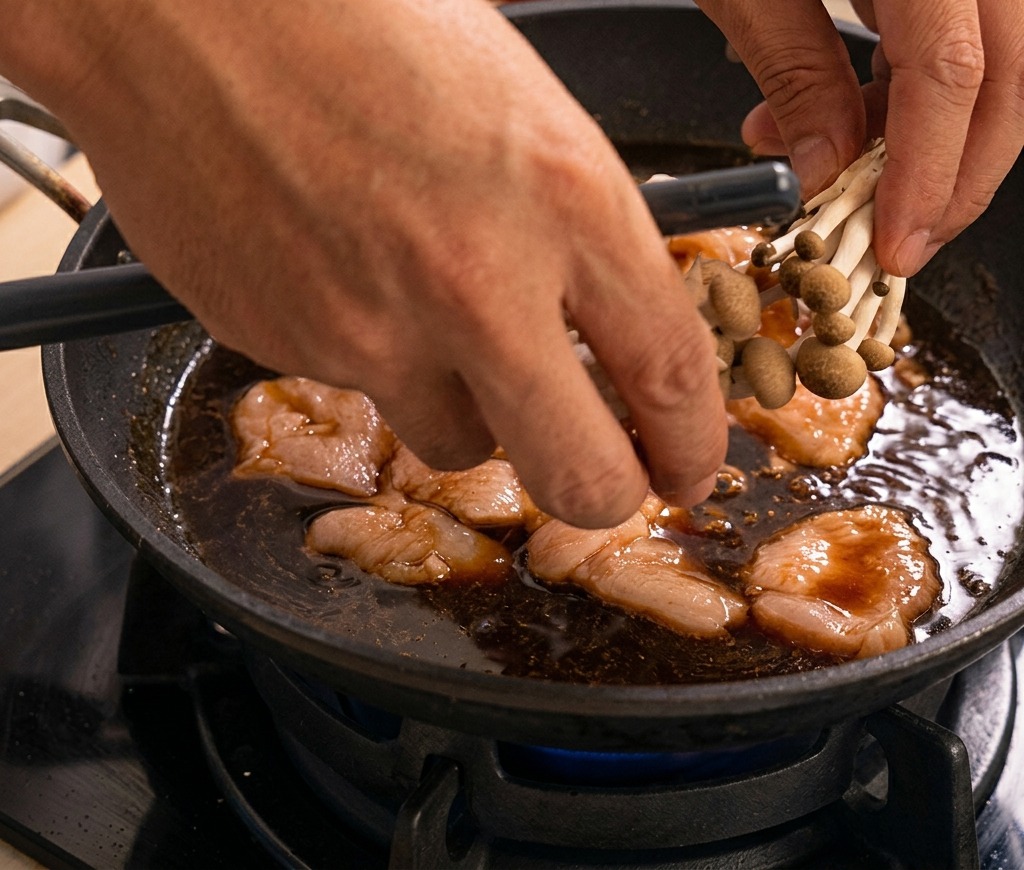 Hände geben frische Shimeji-Pilze zu dem köchelnden Hähnchen in der dunklen Sauce.