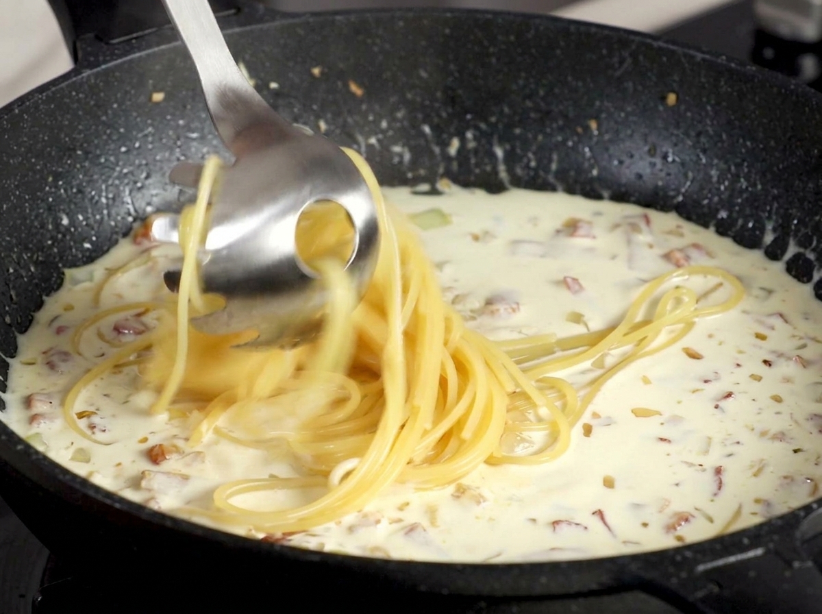 Spaghetti werden mit einem Nudellöffel direkt aus dem Kochwasser in die cremige Specksauce gehoben.