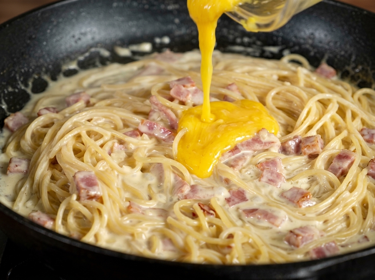 Die gelbe Eigelb-Parmesan-Paste wird über die cremigen Spaghetti in der Pfanne gegeben.