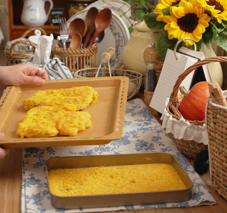 Hände halten ein Backblech mit zwei großen, panierten Schweineschnitzeln in einer warmen Küche.
