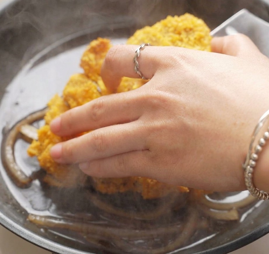 Eine Hand legt Streifen des goldbraun panierten Schweineschnitzels in eine Pfanne mit köchelnden Zwiebelstreifen und dunkler Sauce.
