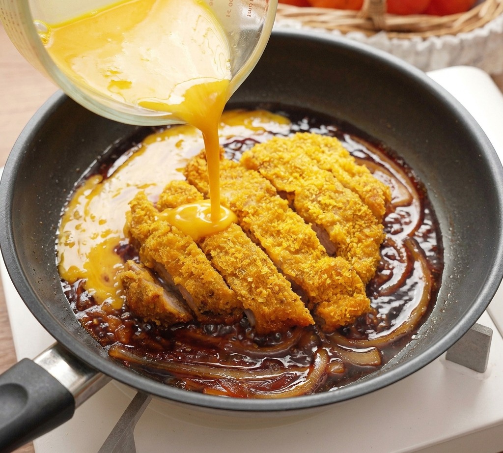 Gelbes verquirltes Ei wird aus einem Glasmessbecher in eine Pfanne mit geschnittenem, gebratenem Schweineschnitzel und Zwiebeln gegossen.