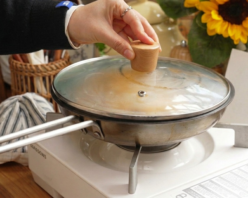 Eine Hand hält den Holzknauf eines Glasdeckels, der auf eine Edelstahlpfanne gesetzt wird, um das Fleisch- und Eiergericht zu dämpfen.