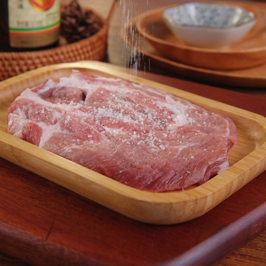 Salz wird aus einer Hand auf ein rohes Schweineschnitzel gestreut, das auf einem Holztablett liegt.
