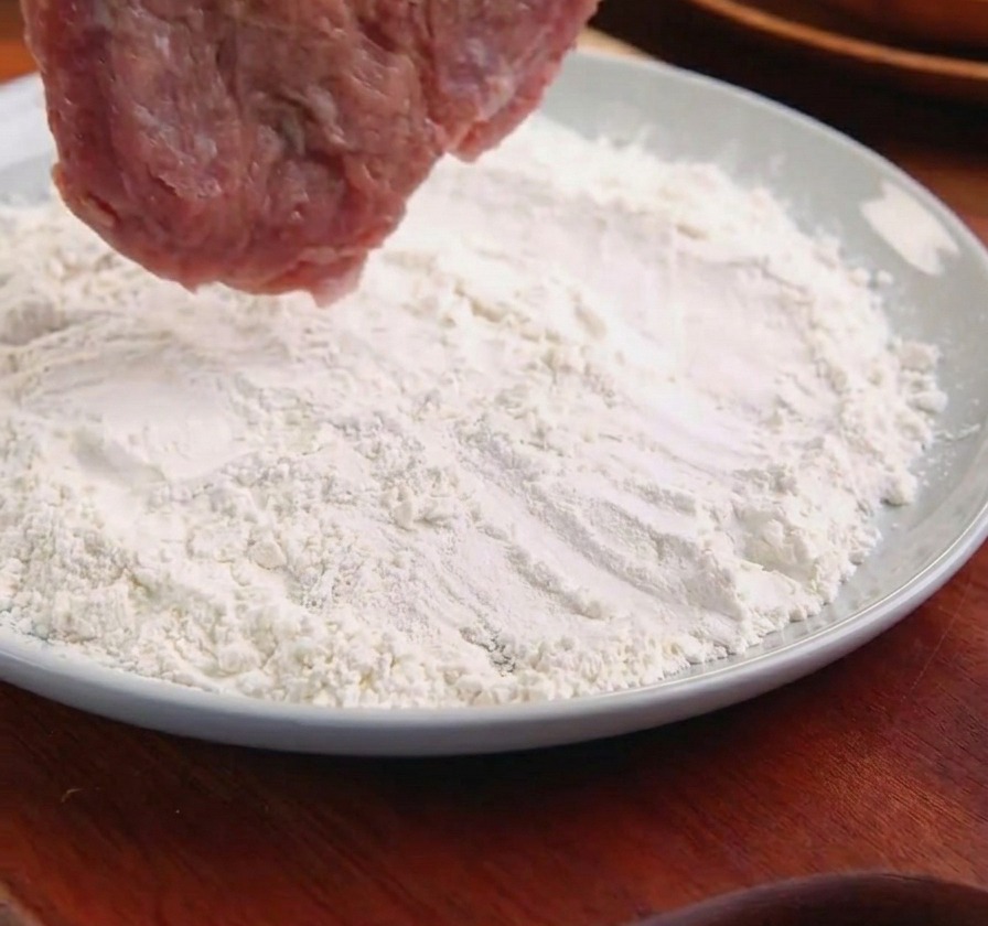 Ein Schweineschnitzel wird mit einer Metallzange in einen Teller mit weißem Mehl gelegt.