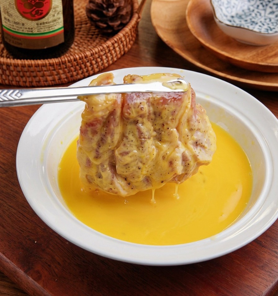 Ein mehliertes Schweineschnitzel wird in eine weiße Schüssel mit verquirltem, gelbem Ei getaucht.