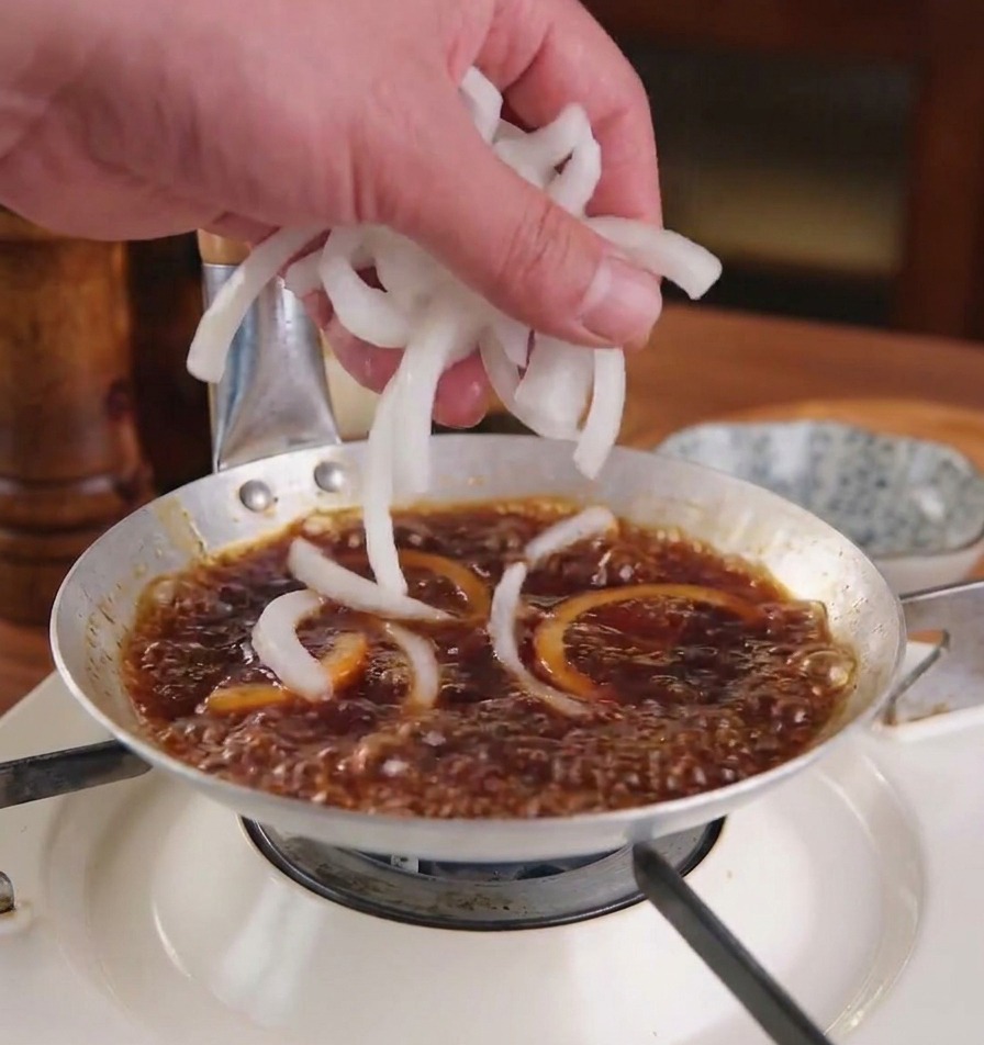 Eine Hand gibt frische weiße Zwiebelscheiben in eine köchelnde, dunkelbraune Sauce in einer flachen Metallpfanne.