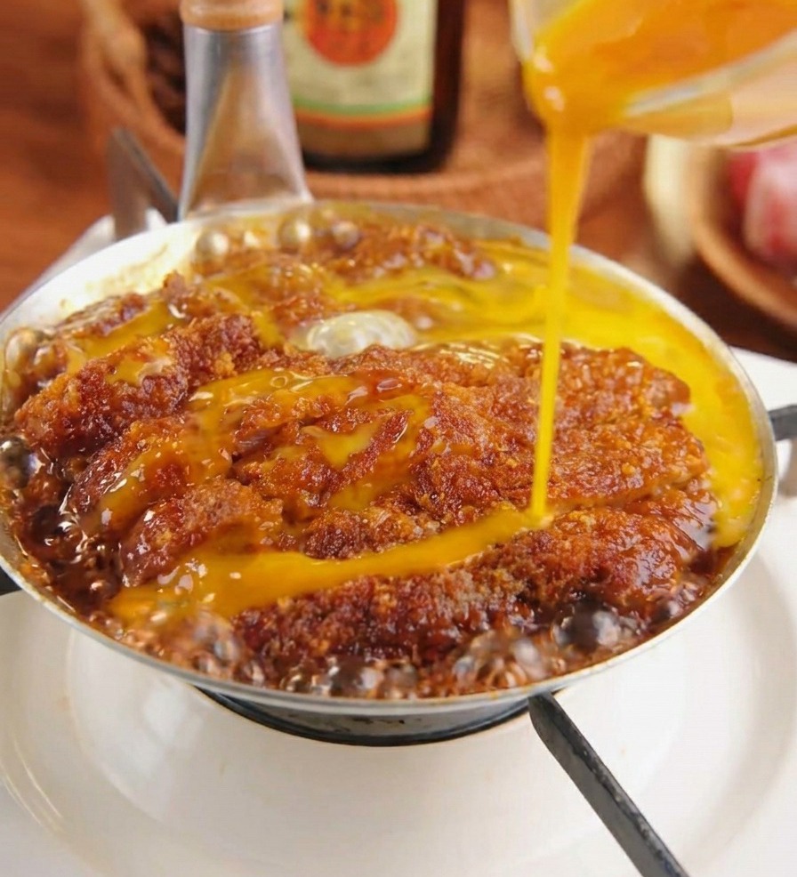 Verquirltes gelbes Ei wird über ein paniertes Schweineschnitzel gegossen, das in einer dunklen Sauce mit Zwiebeln köchelt.