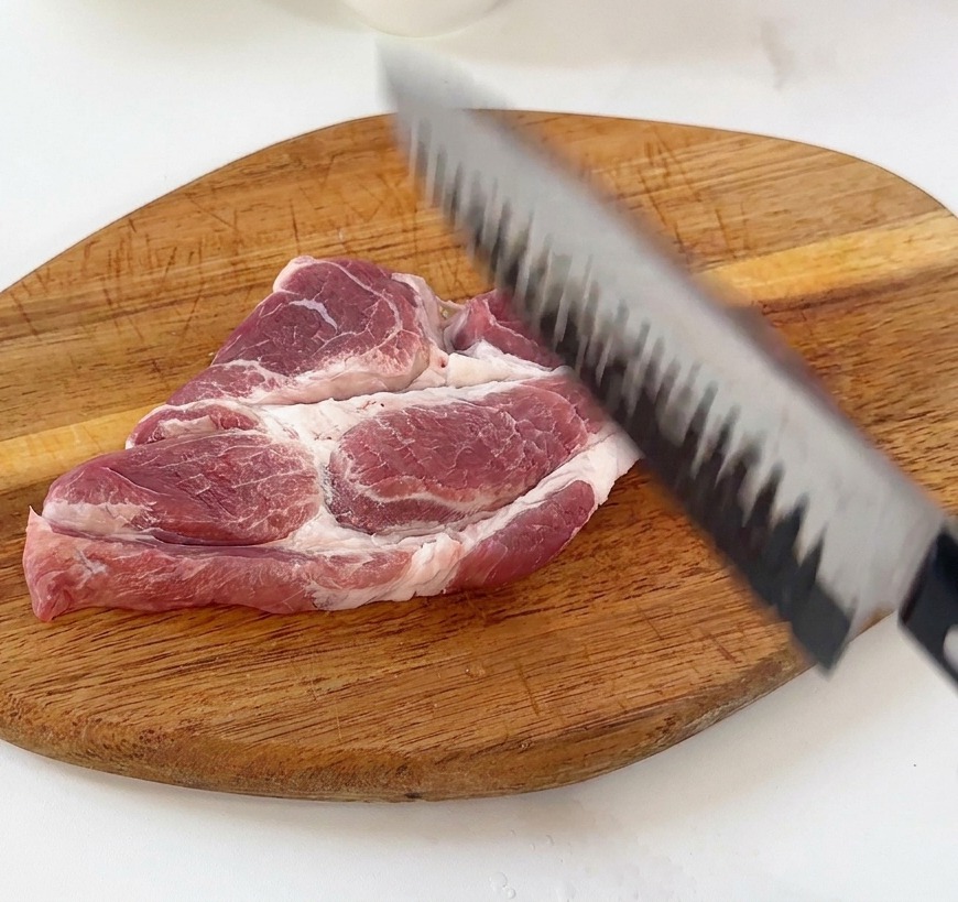 Eine Hand klopft ein rohes Schweineschnitzel auf einem Holzbrett mit dem Messerrücken weich.