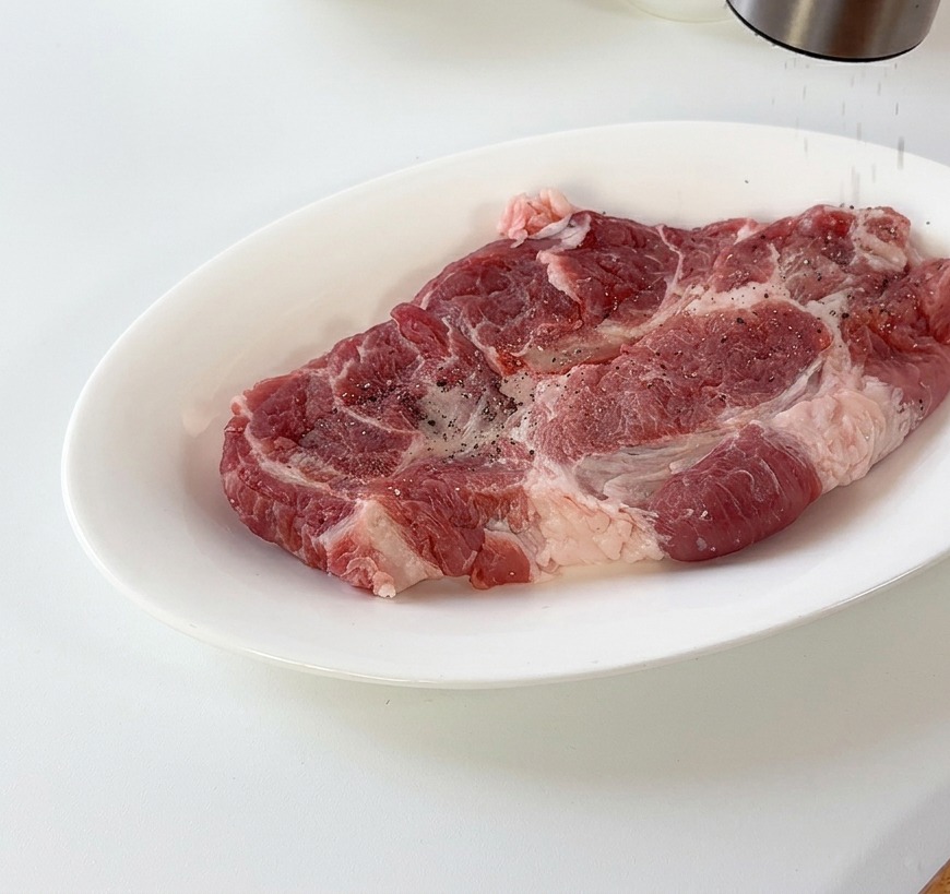 Frischer schwarzer Pfeffer wird über ein rohes Schweineschnitzel auf einem weißen Teller gemahlen.