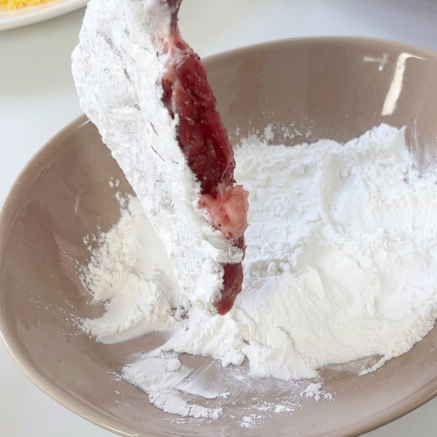 Ein Schweineschnitzel wird in einer beigefarbenen Keramikschüssel mit weißem Mehl bestäubt.