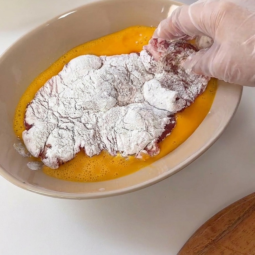Ein mehliertes Schweineschnitzel wird in eine flache Schüssel mit verquirltem gelben Ei getaucht.