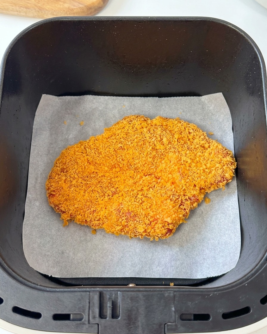 Ein paniertes Schweineschnitzel liegt auf Backpapier im Korb einer schwarzen Heißluftfritteuse.