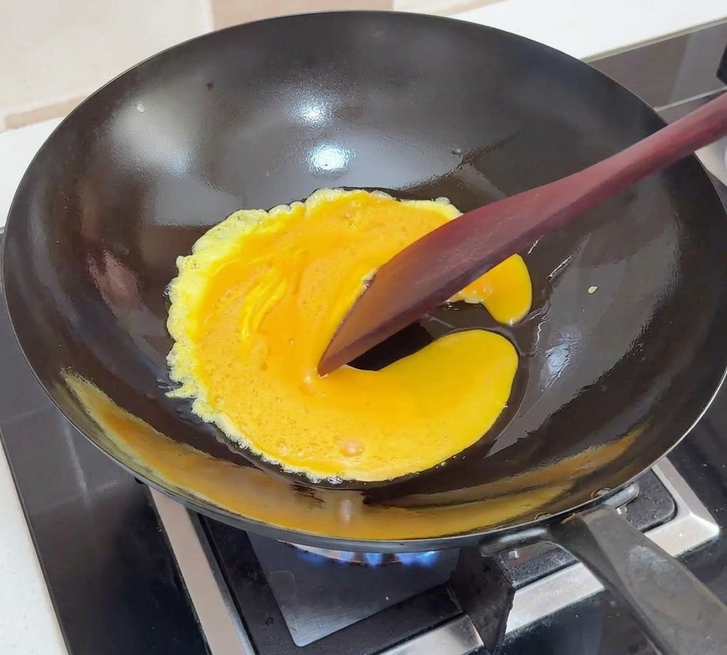 Verquirlte Eier stocken in einem schwarzen Wok und werden mit einem dunklen Pfannenwender sanft zur Mitte geschoben.