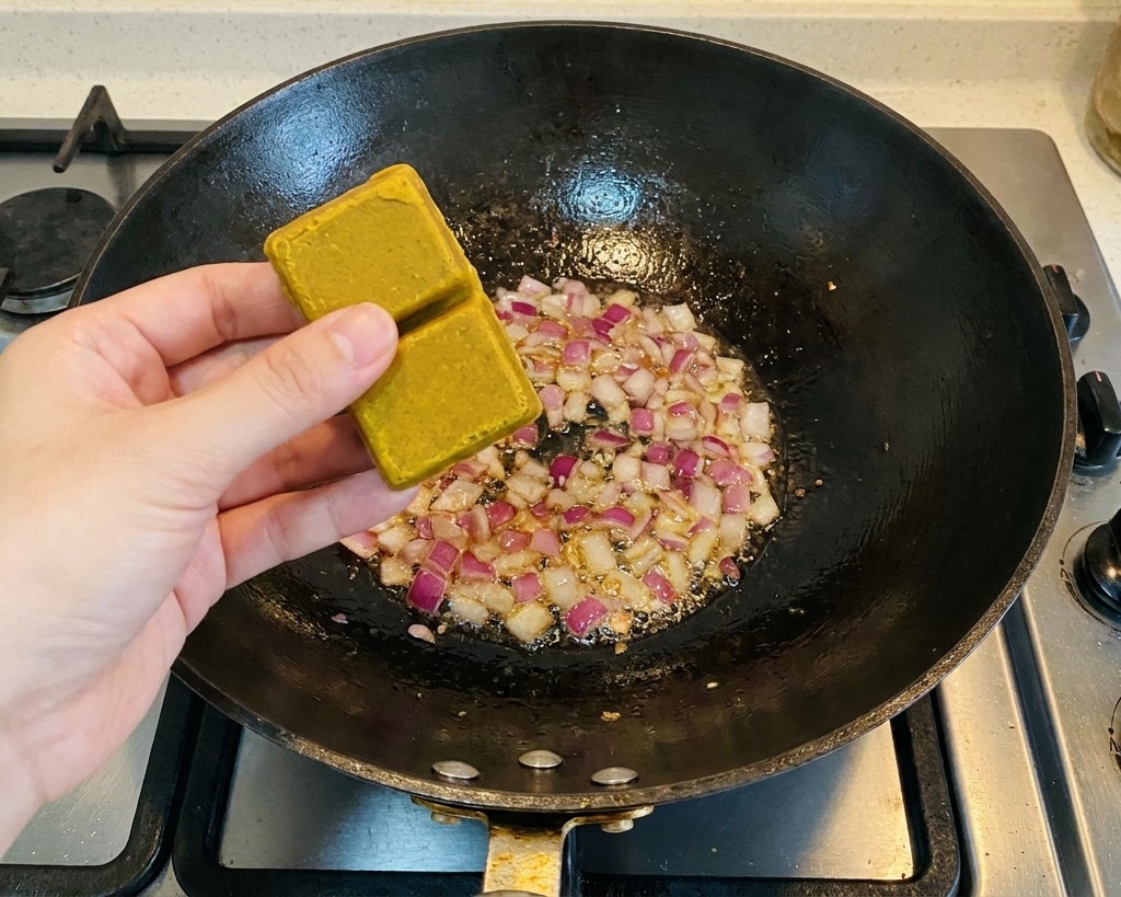 Eine Person hält zwei braune Curry-Würfel über einen Wok mit angedünsteten roten Zwiebelstücken.
