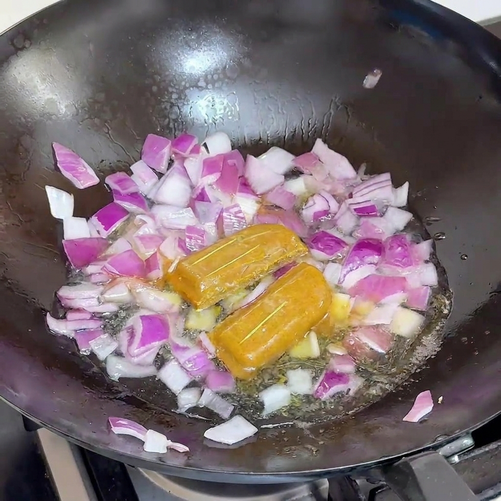 Wasser wird in einen schwarzen Wok gegossen, in dem sich rote Zwiebeln und zwei Curry-Würfel befinden.
