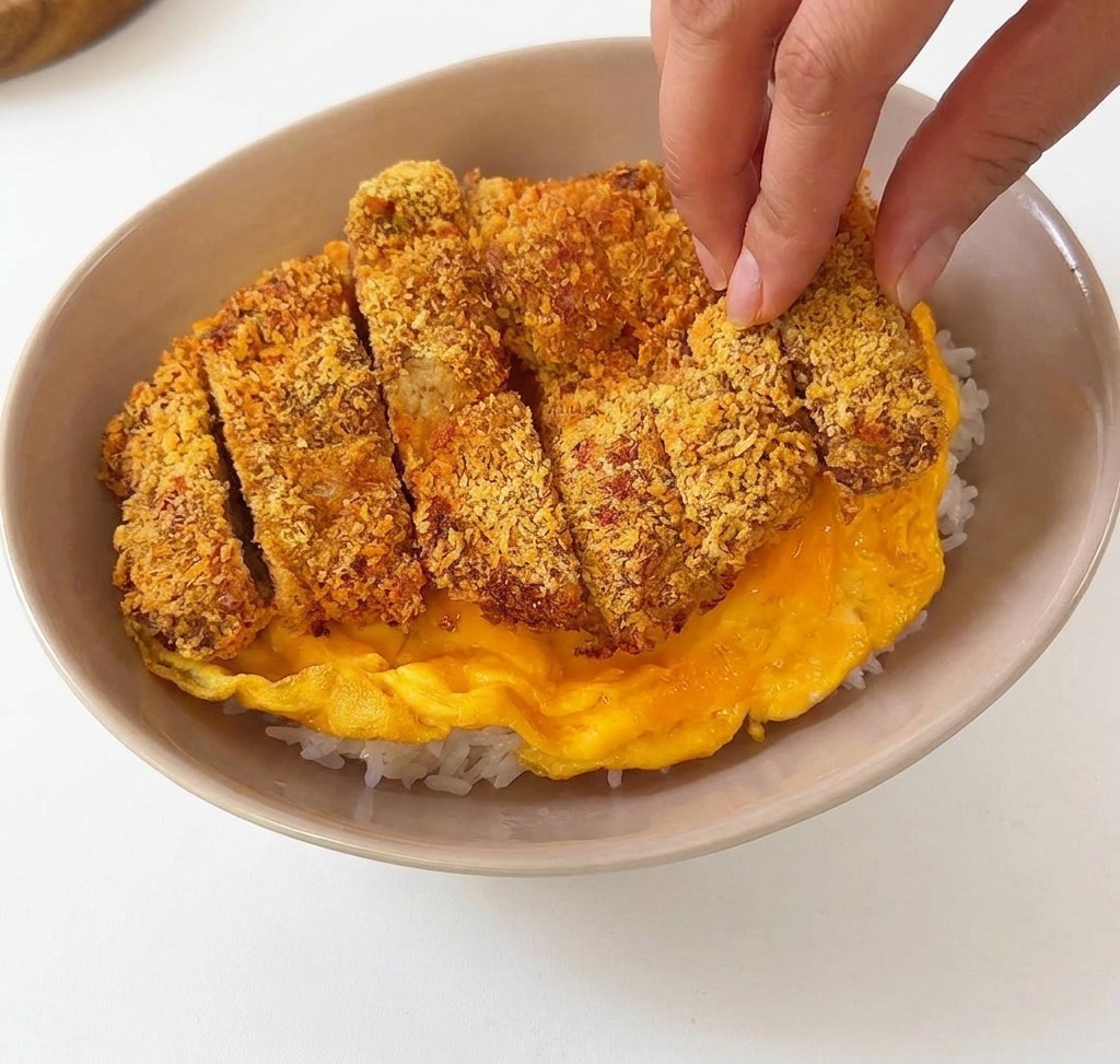 In Streifen geschnittenes paniertes Schweineschnitzel wird auf ein weiches gelbes Omelett in einer beigen Schüssel mit Reis gelegt.