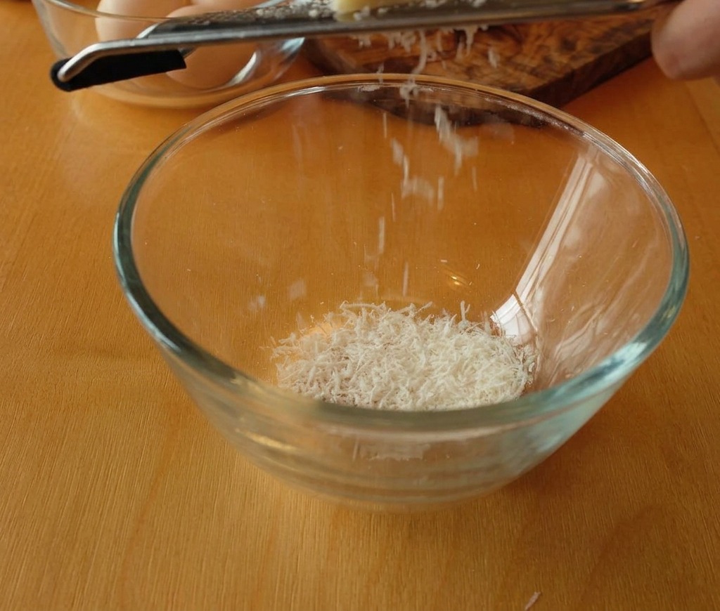 Pecorino Romano wird in eine Glasschale gerieben, sodass feine Käseflocken entstehen.