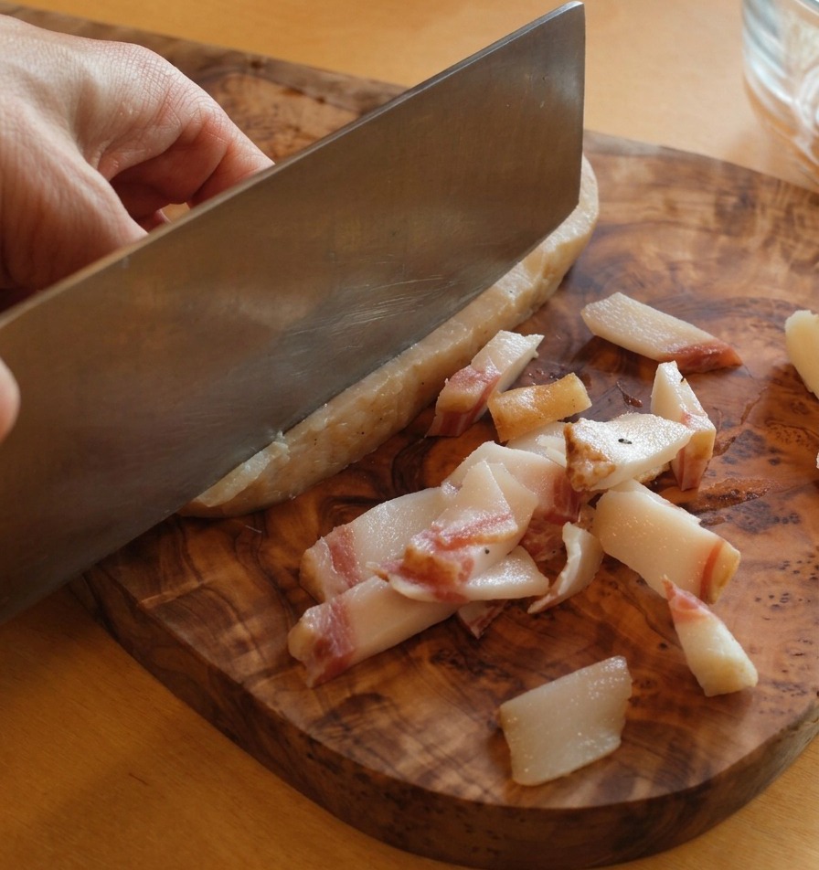 Ein Messer trennt die harte Schwarte vom Guanciale-Stück auf einem Holzbrett.